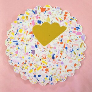 May include: A round, white coaster with a scalloped edge and a gold, cloud-shaped design. The coaster is made of a terrazzo-like material with colorful flecks of pink, blue, yellow, and orange.