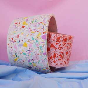May include: Two decorative planters with a terrazzo design. The larger planter is white with colourful flecks, and the smaller one is red with a similar pattern. They are set against a pink backdrop and blue fabric.