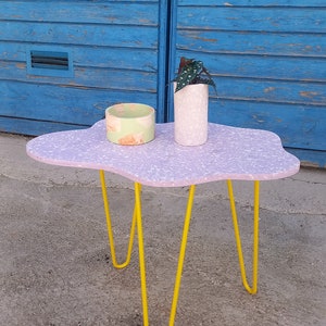 Könnte beinhalten: Ein wolkenförmiger Beistelltisch mit einer Tischplatte im Terrazzo-Stil in Lavendel und Weiß, getragen von leuchtend gelben Haarnadelbeinen. Ein kleiner, runder, hellgrüner und pfirsichfarbener Behälter und ein weißer Topf mit einer Pflanze stehen auf dem Tisch.