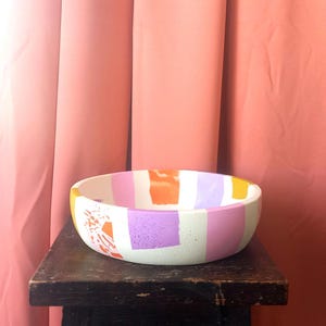 Könnte beinhalten: Eine weiße Keramikschüssel mit einem farbenfrohen geometrischen Design in Orange-, Rosa- und Lilatönen. Die Schüssel steht auf einem dunklen Holzhocker.