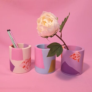 Peut inclure: Trois pots cylindriques en béton aux tons rose, violet et crème, avec des motifs géométriques abstraits. L'un contient des crayons, un autre une fleur, le tout sur fond rose. Les pots mesurent environ 7.6 cm de diamètre.
