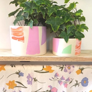 May include: Two white cylindrical planters with abstract designs in pink, orange, and green, holding green ivy plants. The planters sit on a wooden shelf above a floral patterned fabric.