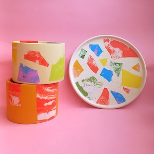 May include: Two colorful ceramic planters with a white background and a colorful ceramic plate with a white background. The planters are stacked on top of each other. The top planter is cream with yellow, green, red, and purple accents. The bottom planter is orange with red and white accents. The plate is white with yellow, green, red, blue, and purple accents.