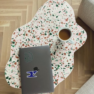 Può includere: Un tavolino da caffè unico, a forma libera, con una superficie in terrazzo maculato bianco, rosa, verde e marrone. Un laptop grigio con adesivi e una tazza bianca con caffè sono sul tavolo. Il tavolo è su un pavimento in legno.
