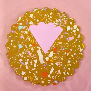 May include: A round, scalloped coaster with a pink, scalloped heart shape in the center. The coaster is made of terrazzo with a yellow base and multicolored flecks.