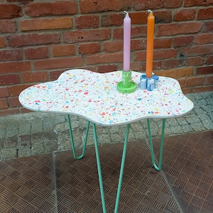 Könnte beinhalten: Ein weißer, wolkenförmiger Tisch mit einer Terrazzo-ähnlichen Oberfläche und grünen Metall-Haarnadelbeinen. Zwei Kerzenständer mit rosa und orangefarbenen Kerzen stehen auf dem Tisch.