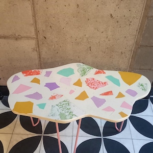 May include: A white cloud-shaped table with a terrazzo-like design featuring pink, purple, green, and yellow accents. The table has four pink hairpin legs and is sitting on a black and white tiled floor.