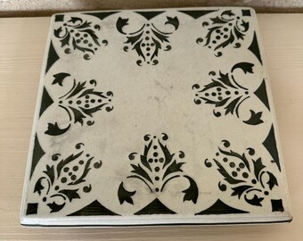 May include: Square, off-white tile with a black floral design. The tile features a repeating pattern of stylized flowers and leaves in each corner and along the edges. The design is symmetrical and creates a decorative border.