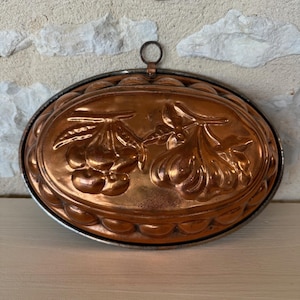May include: An oval copper mold featuring a raised relief design of cherries and pears on the center surface. The mold has a scalloped edge and a small metal ring attached to the top rim for hanging, showcasing a metallic orange-brown finish.