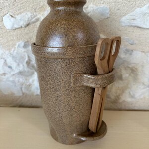 Puede incluir: Un recipiente de cerámica marrón moteado con una parte superior redondeada y un soporte tipo asa. Dos pinzas de madera se colocan en el soporte. El recipiente tiene una superficie texturizada y una base pequeña. El fondo es una pared de piedra clara.