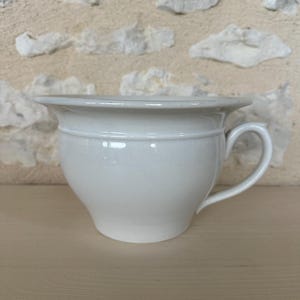 以下が含まれることがあります： 取っ手付きの白い陶器製の便器。ポットは幅広でフレアのリムと丸みを帯びたボディを備えています。背景は明るい色の石壁です。