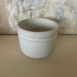 May include: A small, off-white ceramic container with a rounded shape and a slightly textured surface. The container has a simple, classic design, with a subtle ridge near the top. It sits on a light-colored wooden surface, with a textured stone wall in the background.