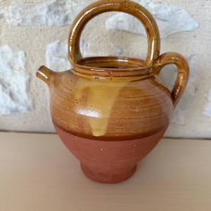 Puede incluir: Una pequeña jarra de cerámica marrón y roja con asa. La jarra tiene un acabado esmaltado y una forma ligeramente redondeada.