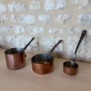Può includere: Tre pentole in rame di dimensioni graduate con interni metallici e lunghi manici in metallo scuro. Ogni pentola presenta un corpo cilindrico con un esterno in rame con patina naturale e un beccuccio sul bordo per versare.