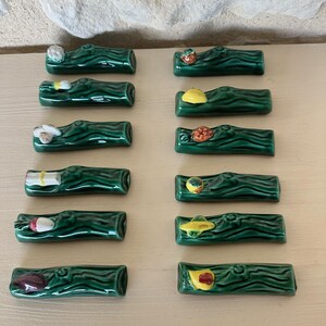Może przedstawiać: Kolekcja ceramicznych podkładek pod pałeczki. Każda podkładka ma kształt ciemnozielonego pnia z innym kolorowym produktem spożywczym na wierzchu, w tym grzybem, truskawką i cytryną. Podkładki są ułożone na jasnobeżowej powierzchni.
