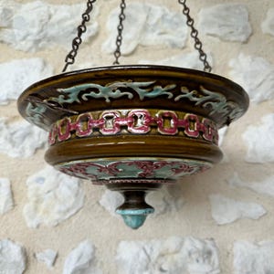 Puede incluir: Una lámpara colgante de cerámica con esmalte marrón, adornada con diseños florales turquesa y rosa, suspendida por una cadena. La lámpara presenta un diseño decorativo en el borde y un remate turquesa. La lámpara está suspendida de una cadena.