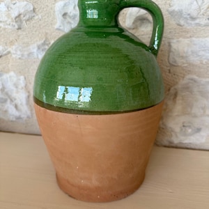 Puede incluir: Jarra de cerámica hecha a mano, de dos tonos. La mitad superior está esmaltada en un verde vibrante, mientras que la mitad inferior es de color terracota natural. Presenta un cuerpo redondeado, un asa y un cuello estrecho. Esta jarra rústica es adecuada para uso decorativo.