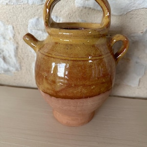 Puede incluir: Una pequeña jarra de cerámica marrón con asa y pico. La jarra tiene un acabado esmaltado y está decorada con una franja marrón alrededor del centro.