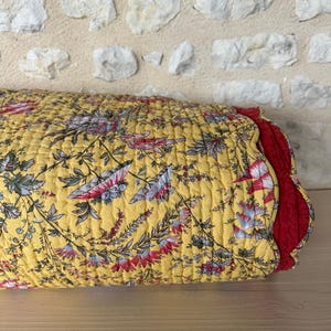Op de afbeelding: Een gele, gewatteerde sprei met bloemenpatroon en rode rand. De sprei heeft een herhalend patroon van roze, blauwe en groene bloemen en bladeren op een gele achtergrond. De sprei is opgevouwen en ligt op een houten oppervlak.