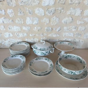 Peut inclure: Ensemble de vaisselle en céramique blanche avec un motif de feuilles vertes. L'ensemble comprend des assiettes, des bols, un plat de service avec couvercle et une assiette de service sur piédestal. Les plats sont disposés sur une surface claire, devant un mur de pierre texturé.