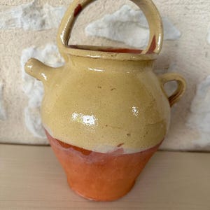 Puede incluir: Una pequeña jarra de cerámica amarilla con un asa marrón y una base marrón. La jarra tiene un pico en un lado y una pequeña asa en el otro.