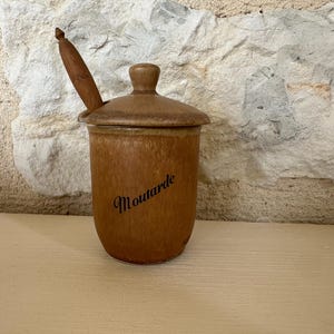 May include: A brown ceramic mustard pot with a lid and a wooden spoon. The word "Moutarde" is written on the side in a cursive font. The pot is sitting on a wooden surface.