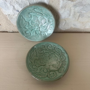 Op de afbeelding: Twee ronde keramische schaaltjes met een licht mintgroen glazuur. Elk schaaltje heeft een verhoogd, reliëf bloemmotief in het midden met gedetailleerde bloemblaadjes en bladeren. De randen van de schaaltjes zijn glad en rond met een glanzende afwerking.