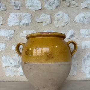 Puede incluir: Una olla de cerámica de dos tonos, con cuerpo redondeado y dos asas pequeñas. La mitad superior es de un amarillo mostaza brillante, mientras que la mitad inferior es beige mate. La olla tiene una abertura ancha y se apoya sobre una superficie de madera contra una pared de piedra.