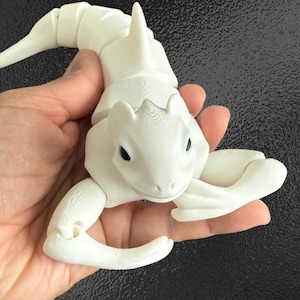 Puede incluir: Una figura de dragón blanco articulado con una aleta dorsal puntiaguda y una larga cola. El dragón tiene pequeños ojos negros y se sostiene en una mano. La figura está hecha de un material impreso en 3D.