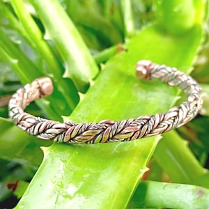 May include: A braided metal cuff bracelet with copper and silver tones. The bracelet is resting on a green aloe vera leaf. The cuff has a textured, woven design and is open at the ends.