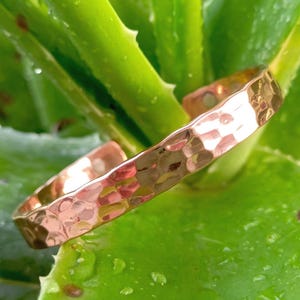 May include: A hammered copper cuff bracelet. The open-ended bracelet has a textured surface and is displayed on a background of green aloe vera leaves. The bracelet is a warm, reddish-brown color.