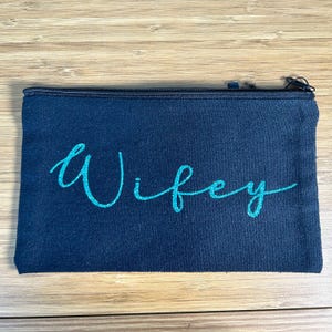 May include: Navy blue canvas zippered pouch with the word "Wifey" in teal script across the front. The pouch has a black zipper and a black zipper pull. The background is a light brown wooden surface.