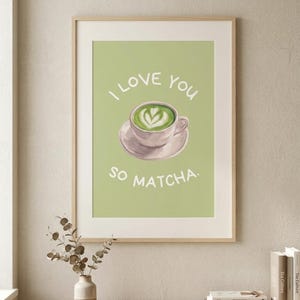 Könnte beinhalten: Ein gerahmter Kunstdruck mit hellgrünem Hintergrund und einer zentralen Illustration einer Tasse Matcha-Latte mit herzförmiger Schaumkunst. Der Text "I LOVE YOU SO MATCHA." ist in Weiß über und unter der Tasse abgebildet.