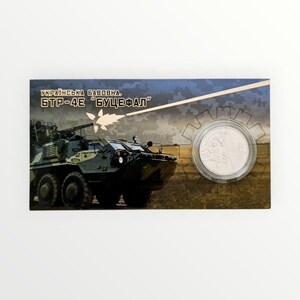 May include: A rectangular display card featuring a green and black camouflage armored vehicle on the left and a circular silver coin encased in a clear plastic capsule on the right. The text "УКРАЇНСЬКА БАВОВНА. БТР-4Е "БУЦЕФАЛ"" is printed in gold.
