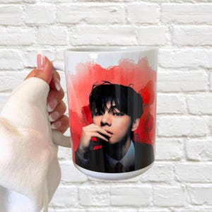May include: White ceramic mug featuring a portrait of a person with black hair and a suit, set against a red watercolor background. The mug has a white handle and is held against a white brick wall.
