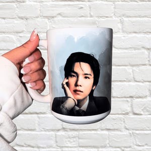 May include: White ceramic mug featuring a portrait of a person with black hair and a dark suit. The background has a light blue watercolor effect. The mug is held by a person with a white sleeve.