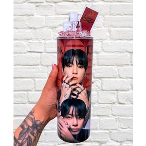 May include: A red and black tumbler featuring two images of a person. The tumbler has a clear lid with decorative ice-like accents and a small card. The background is a white brick wall.