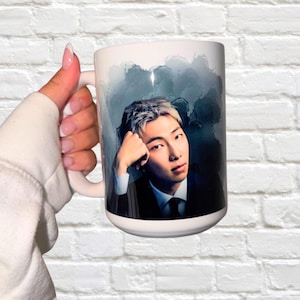 May include: White ceramic mug featuring a portrait of a man with silver hair, wearing a suit and tie, against a blue and gray watercolor background. The mug is held by a person wearing a white sweatshirt.
