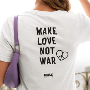 Könnte beinhalten: Weißes T-Shirt mit dem Text "MAKE LOVE NOT WAR" in schwarzen Buchstaben und einem Peace-Zeichen-Herz-Grafik. Der Markenname "NIISE clothing" ist darunter aufgedruckt. Eine Person trägt das T-Shirt und eine beige Baseballkappe.