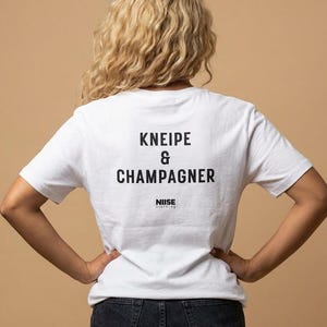 以下が含まれることがあります： 黒字で「KNEIPE & CHAMPAGNER」とプリントされた白いTシャツ。Tシャツにはブランド名「NIISE」もプリントされています。モデルは濃い色のデニムジーンズを着用しています。