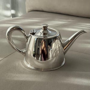 Puede incluir: Una tetera de tono plateado pulido con cuerpo redondeado, asa curva, tapa con bisagra y un pequeño pomo, además de un pico vertedor cónico. La superficie metálica es altamente reflectante y muestra un claro reflejo distorsionado.