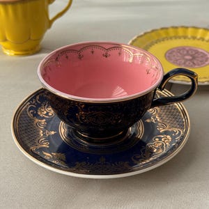 Puede incluir: Un juego de taza y plato de color azul marino oscuro con intrincados patrones de filigrana dorada y un interior rosa suave. La taza tiene un borde dorado con un diseño festoneado. Una taza amarilla y un plato amarillo son visibles al lado.