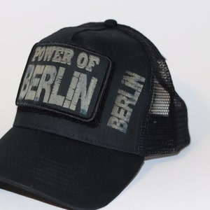 Könnte beinhalten: Schwarze Trucker-Kappe mit schwarzem Mesh-Rücken. Die Vorderseite der Kappe hat einen schwarzen Patch mit den Worten "POWER OF BERLIN" in weißer Schrift. Die Seite der Kappe hat das Wort "BERLIN" in weißer Schrift.