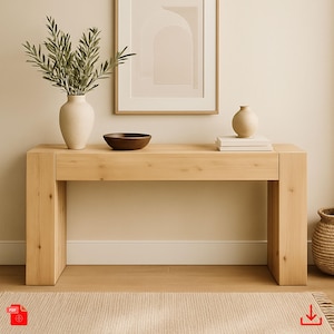 Puede incluir: Una mesa consola de madera clara con un acabado natural. La mesa tiene una tapa rectangular y dos patas cuadradas. Hay un cuenco, un jarrón con vegetación y una pila de libros sobre la mesa.