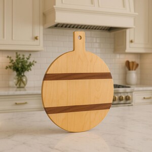 Op de afbeelding: Een ronde houten snijplank met een handvat. De plank is licht van kleur met twee donkerbruine banden. Het handvat heeft een gat om op te hangen. De plank wordt gepresenteerd op een wit aanrecht in een keuken.