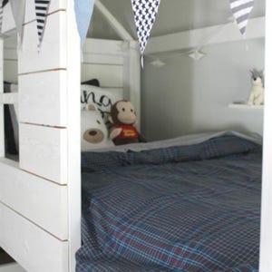 May include: A white wooden bed frame with a house-like structure, decorated with fabric bunting. The bed is made with a blue plaid comforter and pillows. Stuffed animals, including a bear and monkey, are on the bed.