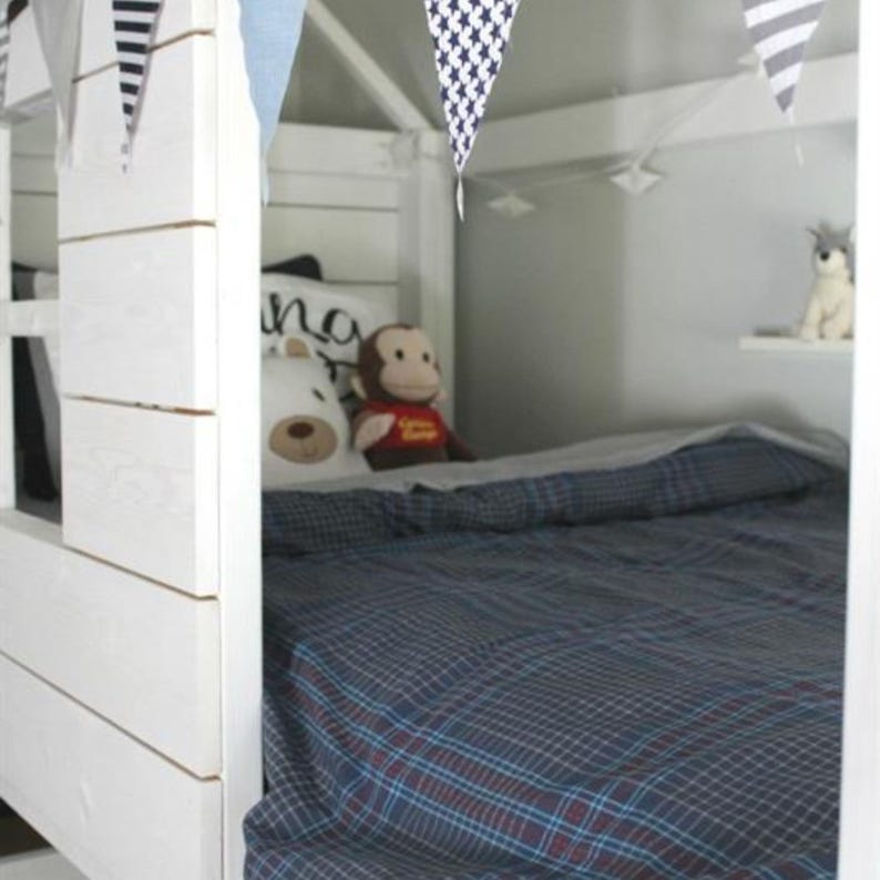 May include: A white wooden bed frame with a house-like structure, decorated with fabric bunting. The bed is made with a blue plaid comforter and pillows. Stuffed animals, including a bear and monkey, are on the bed.