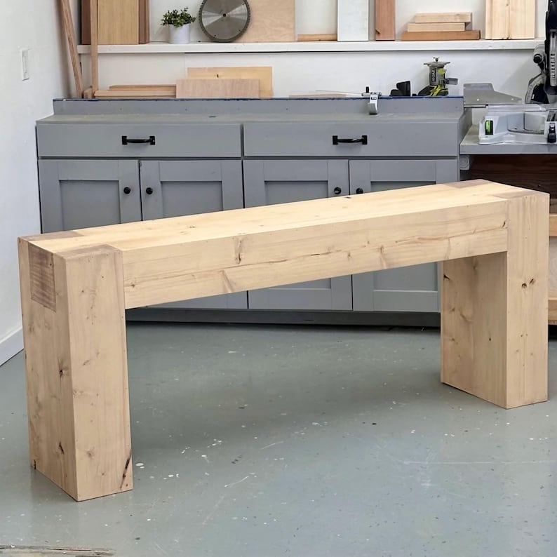 May include: A light wood console table with a simple design. The table has a rectangular top and two square legs. The table is made of unfinished wood and has a natural wood grain.