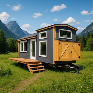 May include: A mobile tiny house, painted grey with natural wood features, is positioned on a grassy field. The house has a small deck, steps, and several windows. Mountains and a blue sky with clouds are in the background.