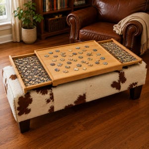 May include: A wooden puzzle table with a cowhide patterned ottoman base. The table has three sections, with two sliding trays filled with puzzle pieces and a central area with a partially assembled puzzle. The table is made of light-coloured wood.
